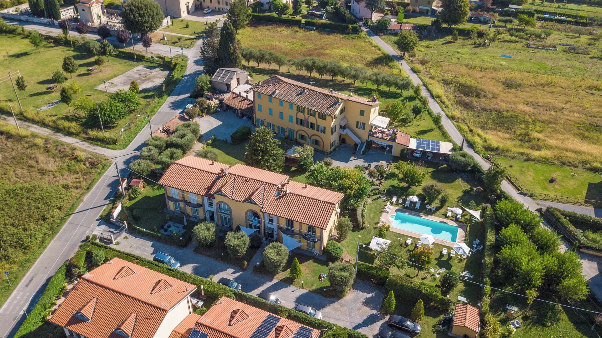 A large, yellow building with a swimming pool, surrounded by greenery and a road.