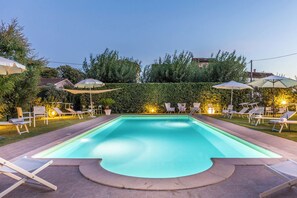 A rectangular swimming pool with a curved edge, surrounded by lounge chairs and umbrellas, set against a backdrop of a hedge and trees. - New Listing (Lucca)