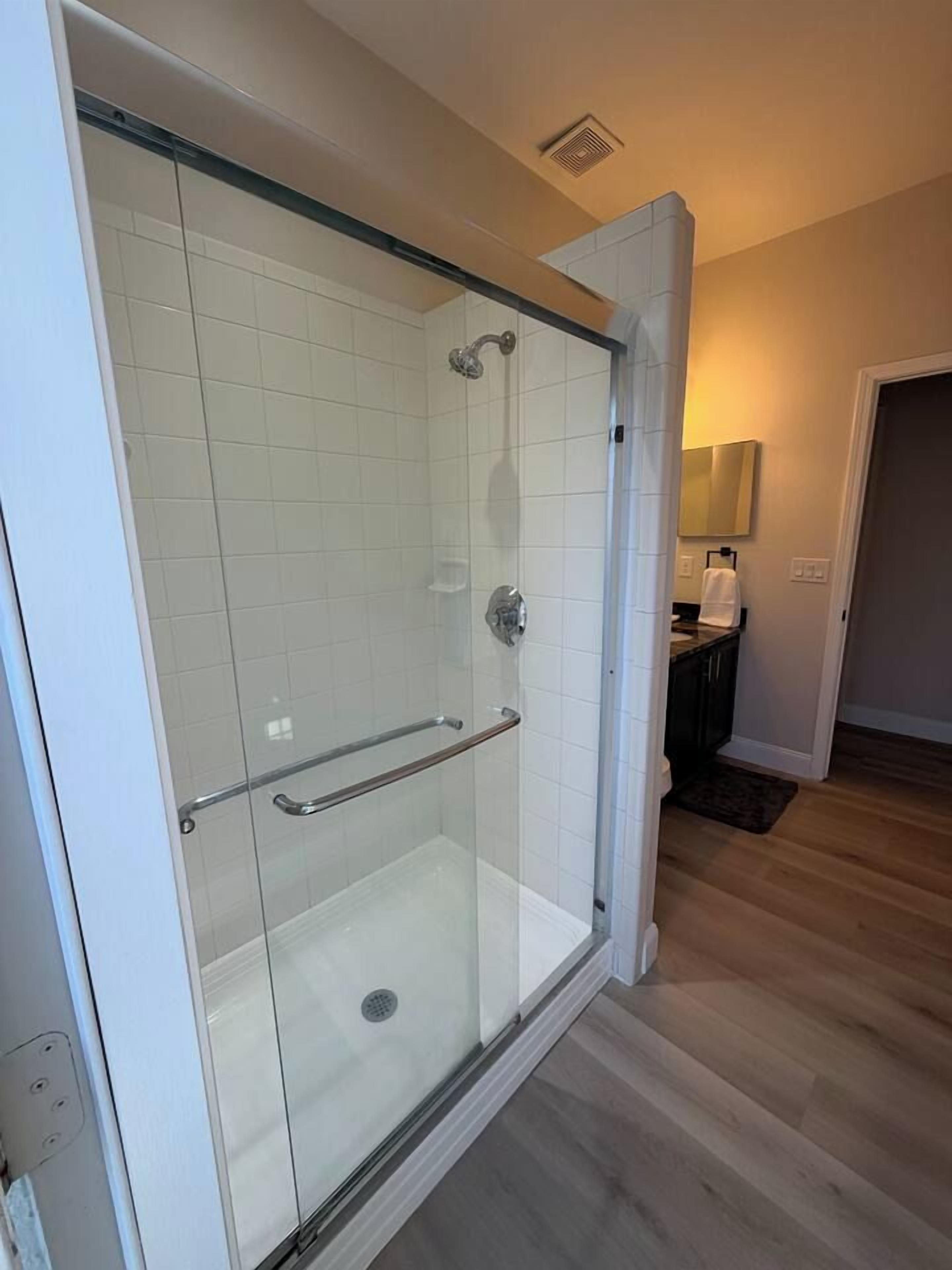 Ducha con cerramiento de vidrio, barra de apoyo, pared con azulejos y piso de madera.