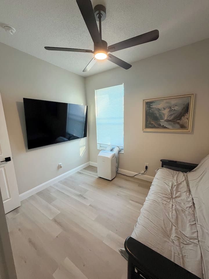 Una habitación con ventilador de techo, televisión montada en la pared, ventana con persianas, un cuadro en la pared y un sofá blanco.