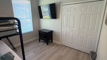 Un dormitorio con una cama litera, ventilador de techo, una mesita pequeña, televisor montado en la pared y una puerta blanca.