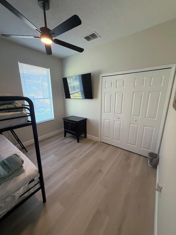 A bedroom with a bunk bed, a ceiling fan, a small table, a TV mounted on the wall, and a white door.