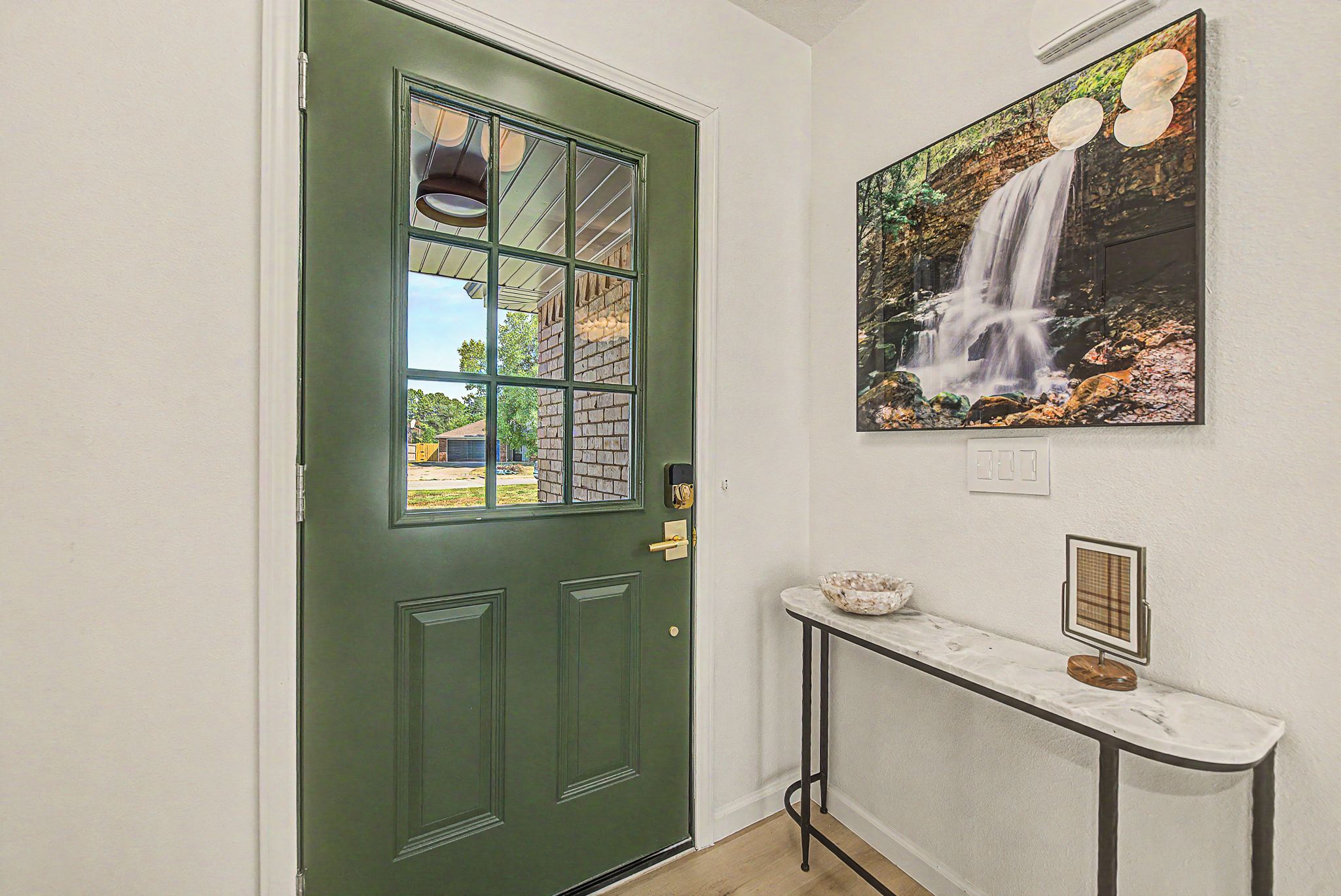 A green door with a window, a wall-mounted picture of a waterfall, and a small table with a bowl and a framed picture.