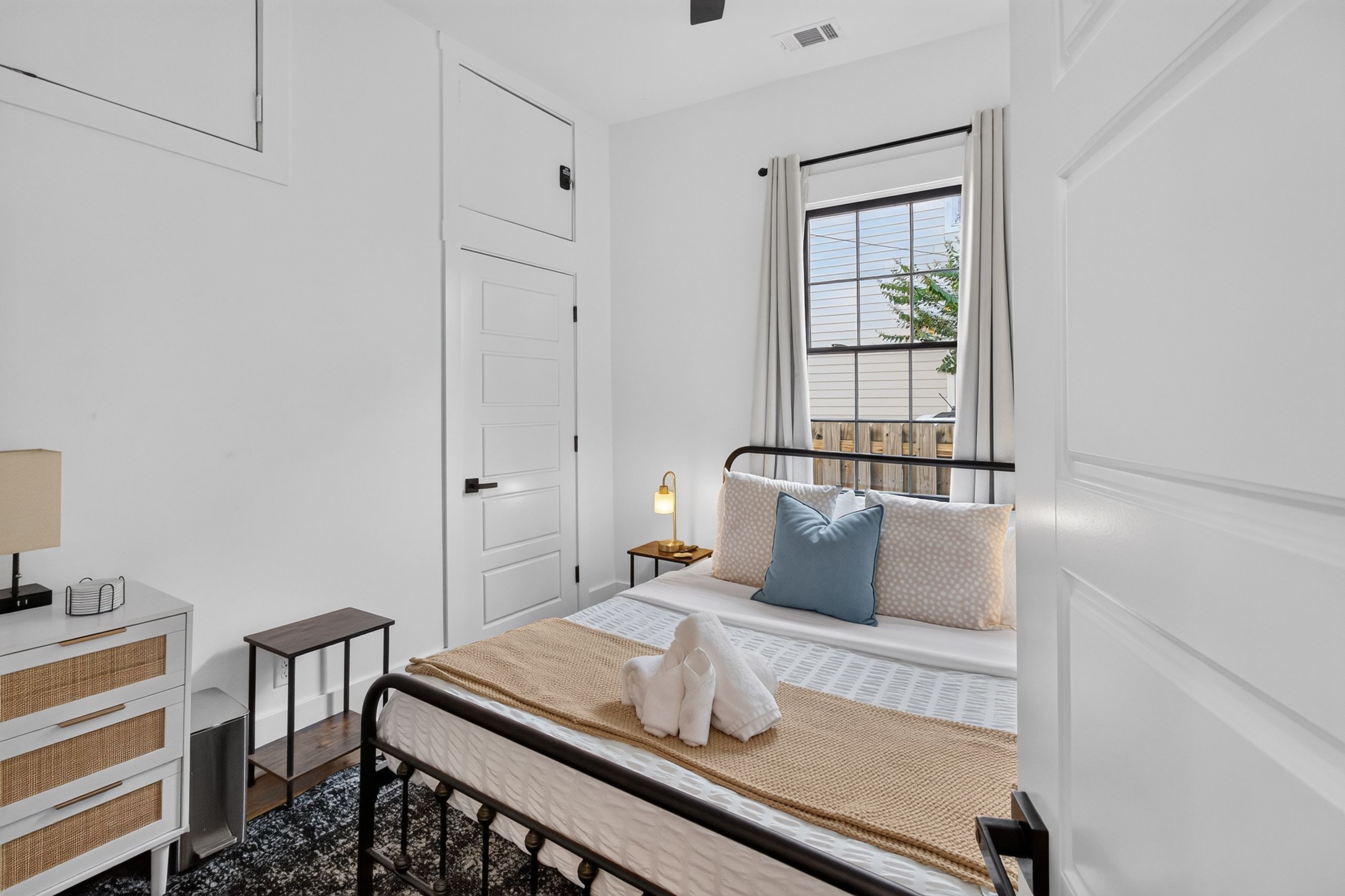 A bedroom with a bed, bedside table, lamp, and a window with a view of the outside.