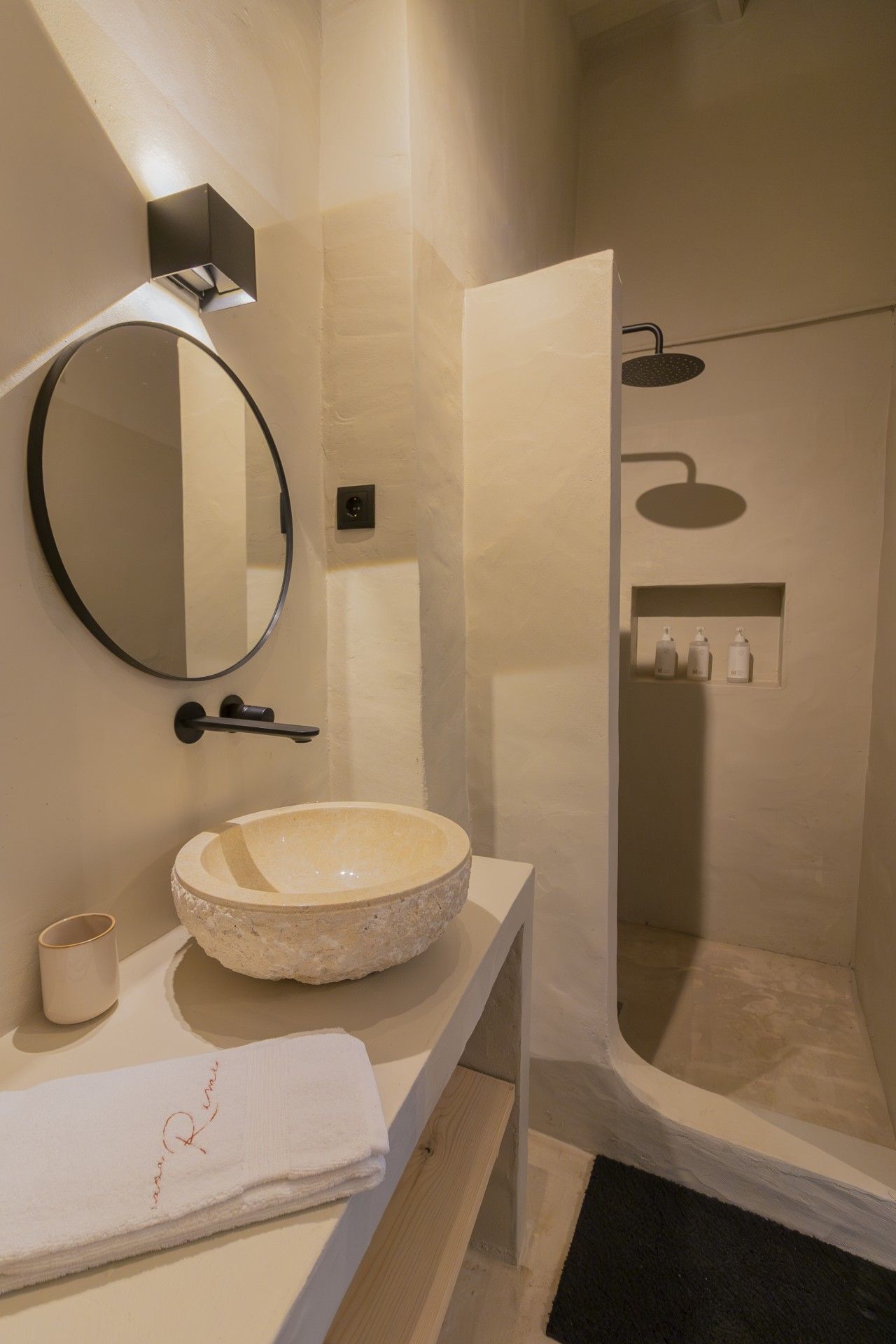 Un bagno moderno con un lavandino in marmo rotondo, uno specchio fissato alla parete, un soffione doccia manuale e un piccolo ripiano con articoli da toilette.