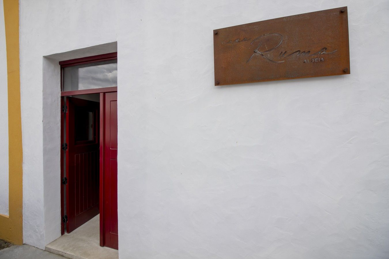 Een rode deur met daarboven een klein raam, een witte muur en een metalen plaquette met de naam 'Rama' en 'ALGEIA'.