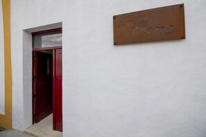 Una puerta roja con un pequeño ventanal encima, una pared blanca y una placa metálica con los nombres 'Rama' y 'ALGEIA'.