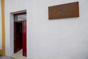 Eine rote Tür mit einem kleinen Fenster darüber, eine weiße Wand und eine Metallplakette mit den Namen 'Rama' und 'ALGEIA'.