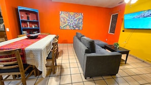 Una sala de estar con un sofá, una mesa de comedor con sillas, una estantería y un cuadro abstracto y colorido en la pared.