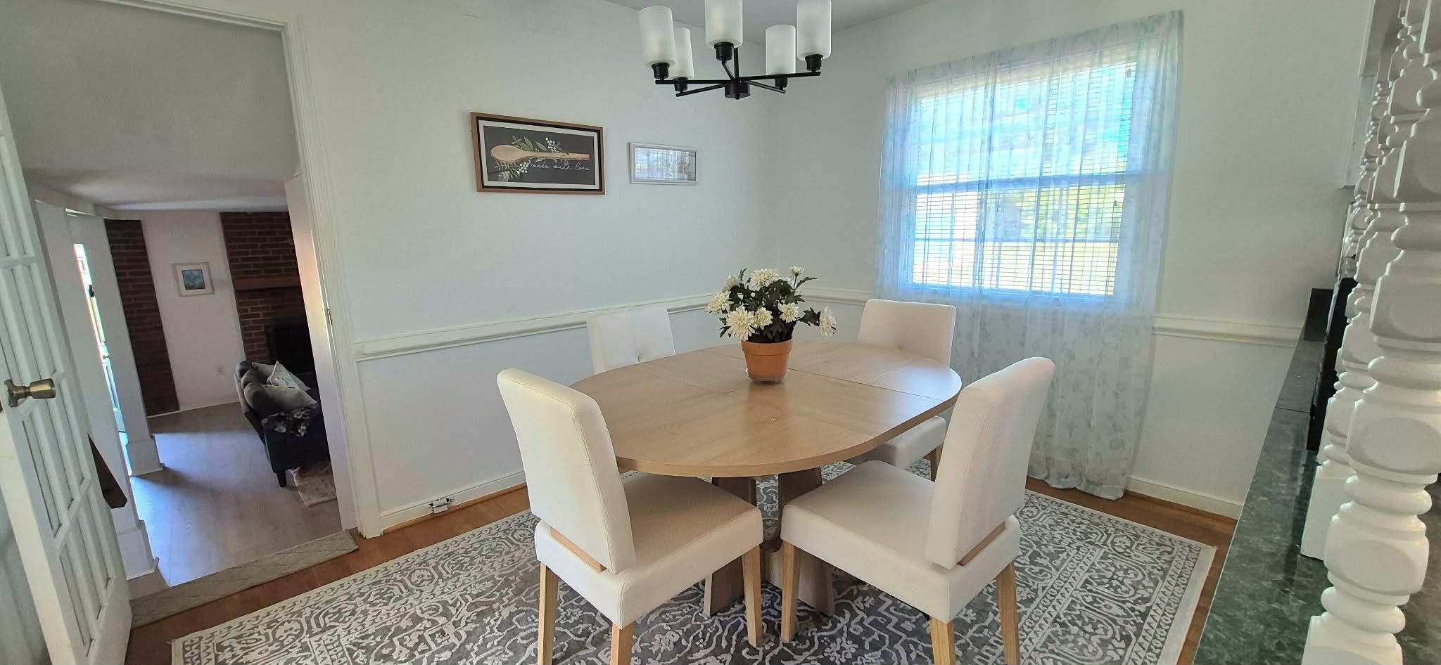 A dining area with a round table, four chairs, a chandelier, and a window with curtains.