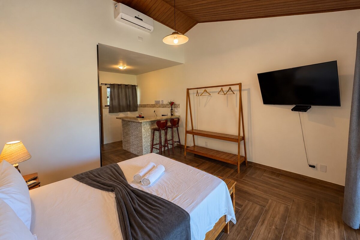 Une chambre d’hôtel avec un lit, une télévision, un plafond en bois et, au fond, une petite cuisine.