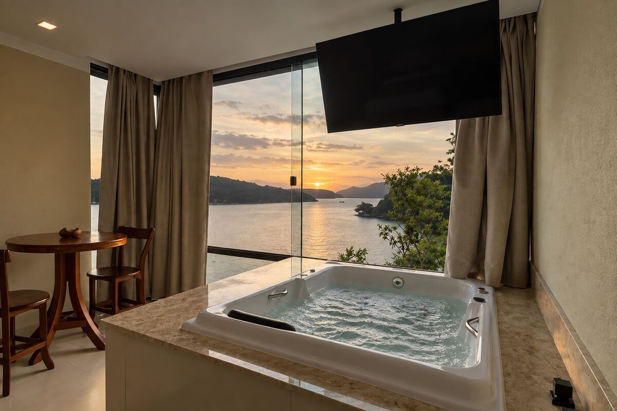 Une salle de bain moderne avec une grande baignoire, une petite table et une chaise, donnant sur une étendue d’eau au coucher du soleil.