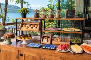 Un buffet proposant une variété de plats, notamment des pâtisseries, des fruits et des desserts, présentés sur des étagères en bois, avec une vue sur la mer en arrière-plan.