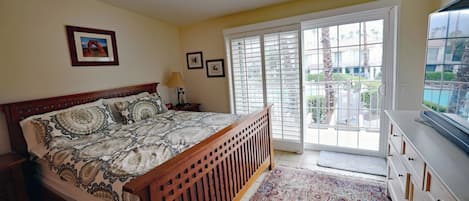 A bedroom with a bed, a nightstand, a lamp, a window with blinds, and a view of a pool and buildings outside.