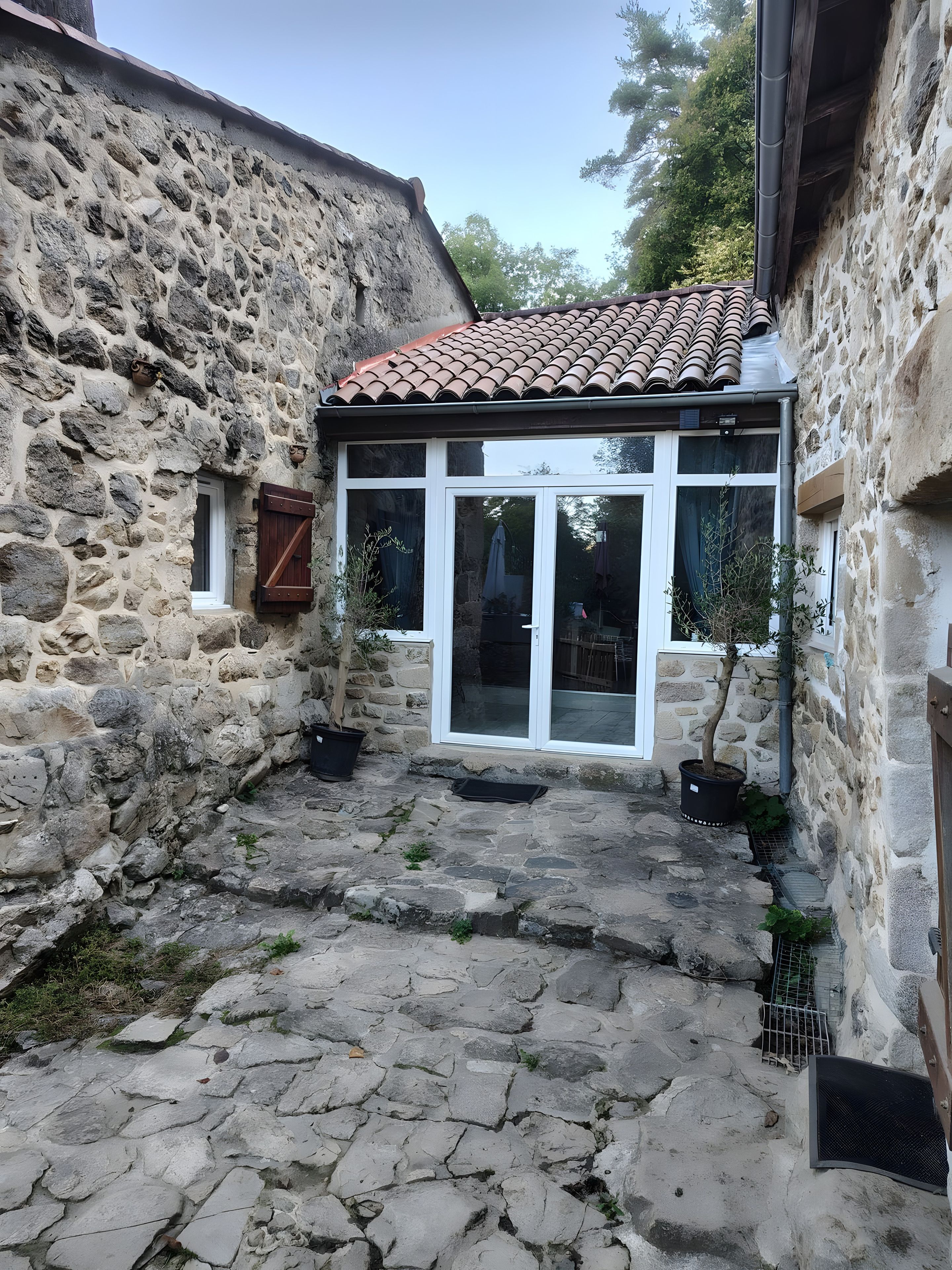 Un bâtiment en pierre avec un toit en tuiles, une porte d’entrée en verre et un pot de fleurs.