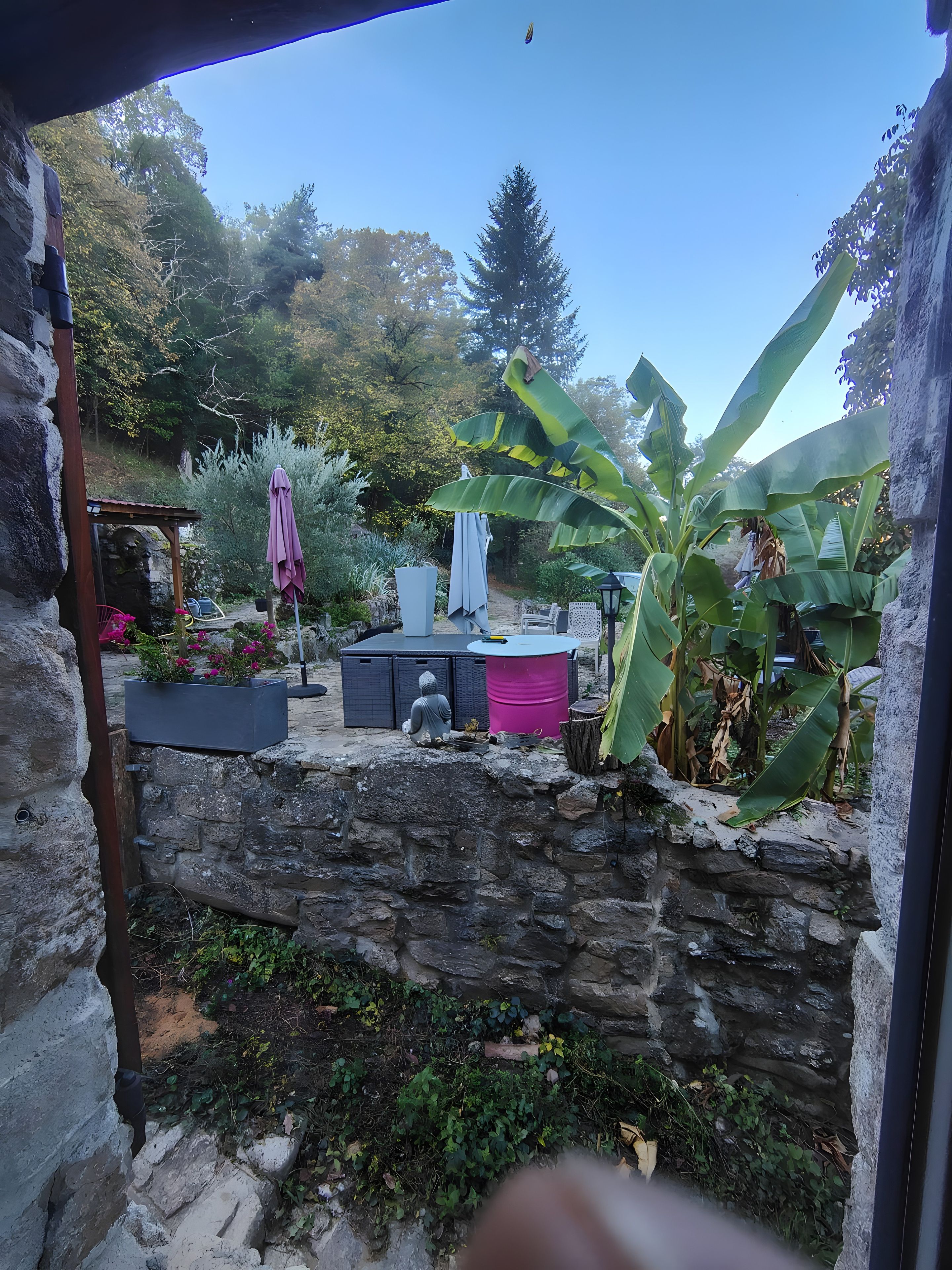 A stone wall with a garden area, including a seating area with a table and chairs, a pink barrel, and a statue.