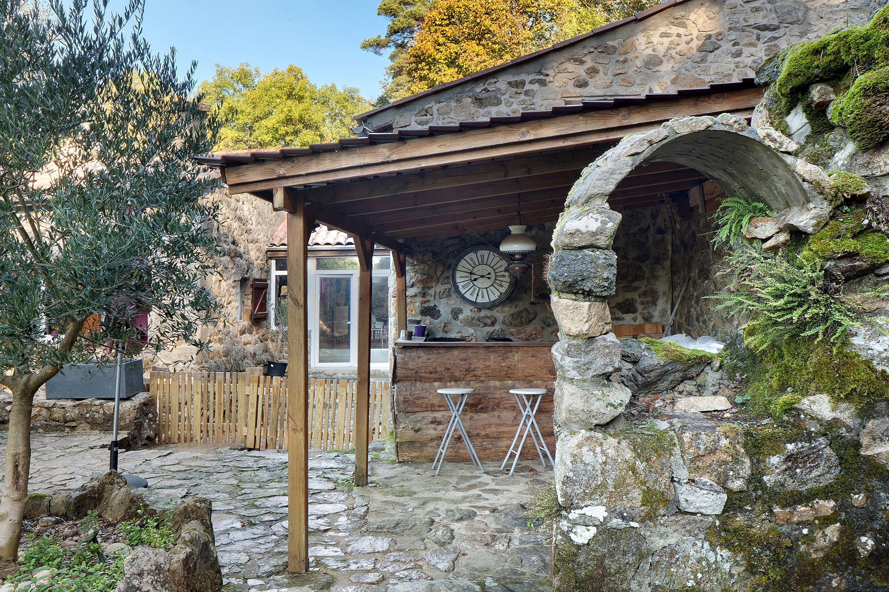 Un edificio rústico de piedra con una pérgola de madera, paredes de piedra y un reloj de piedra.