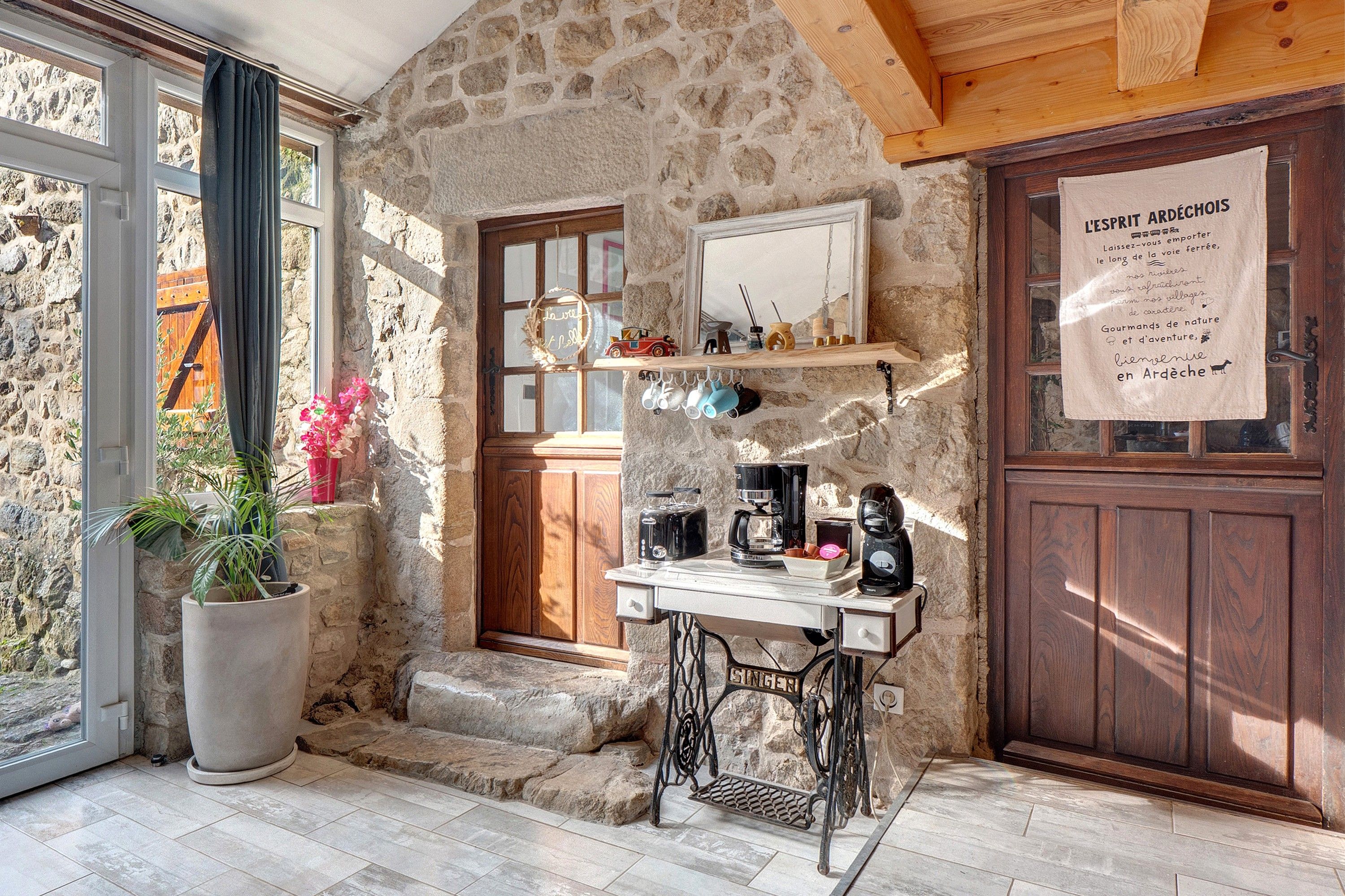 Una habitación rústica con paredes de piedra, una puerta de madera, una ventana con cortinas, una máquina de coser y un cartel en francés.