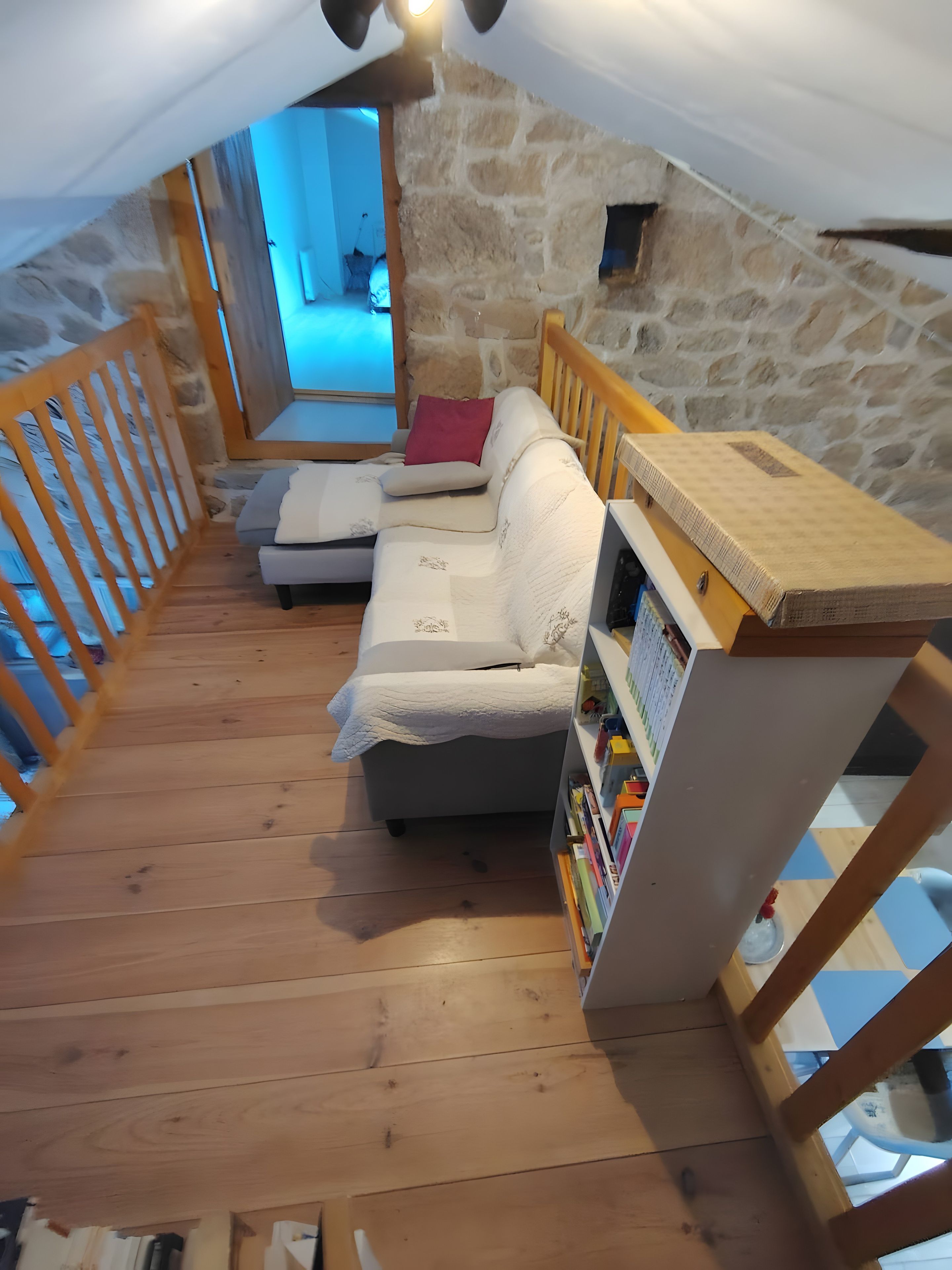 Une pièce mansardée avec un plancher en bois, un canapé blanc, une bibliothèque et un mur en pierre.