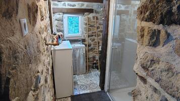 Un couloir étroit aux murs de pierre menant à une salle de bain équipée d’une douche et d’un lavabo.
