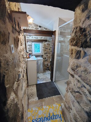 Ein schmaler Gang mit Steinwänden, der zu einem Badezimmer mit Dusche und Waschbecken führt.