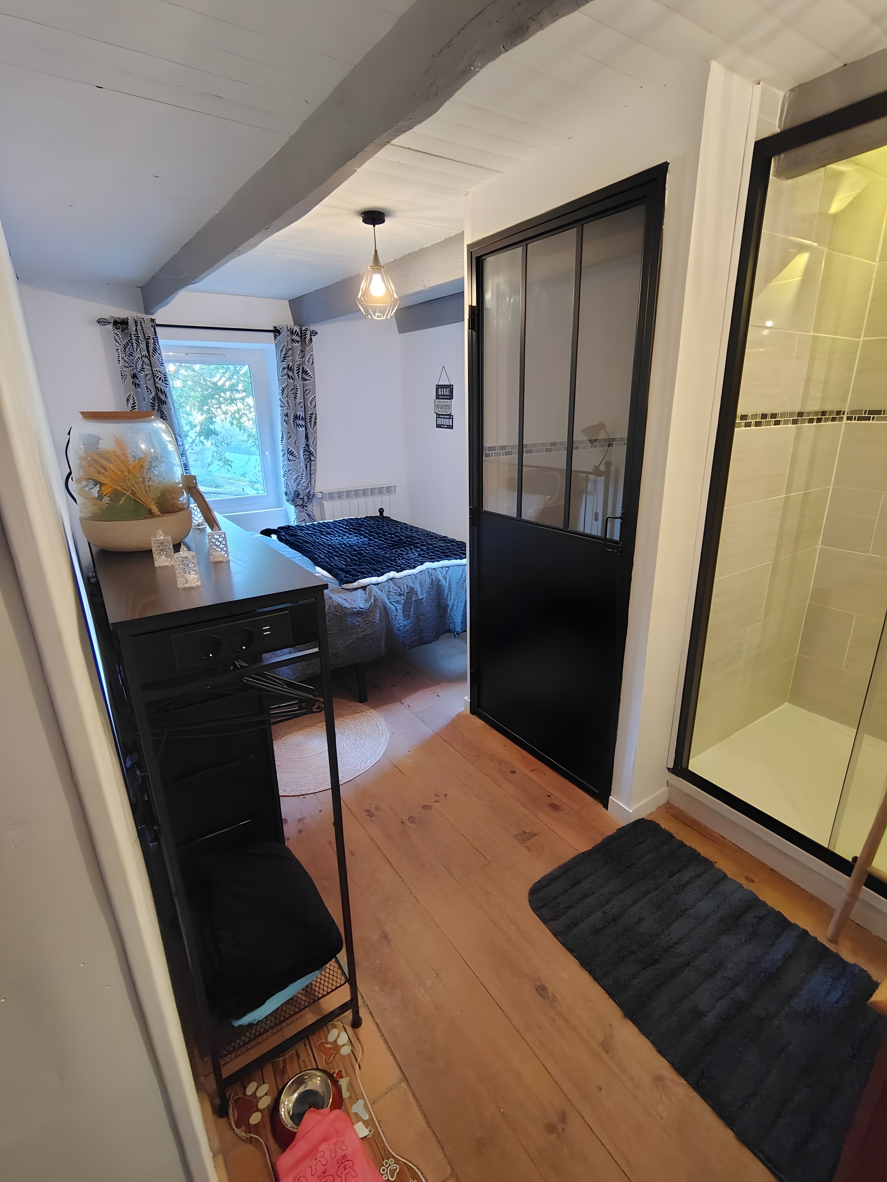 Une pièce de taille modeste comprenant un lit, une petite table avec un bol et des plantes séchées, un meuble noir et une porte coulissante en verre donnant sur un espace douche.
