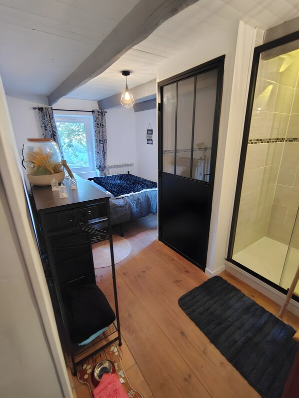 Habitación compacta con cama, una mesita con un tazón y plantas secas, un mueble negro y una puerta corrediza de vidrio que da a una zona de ducha.