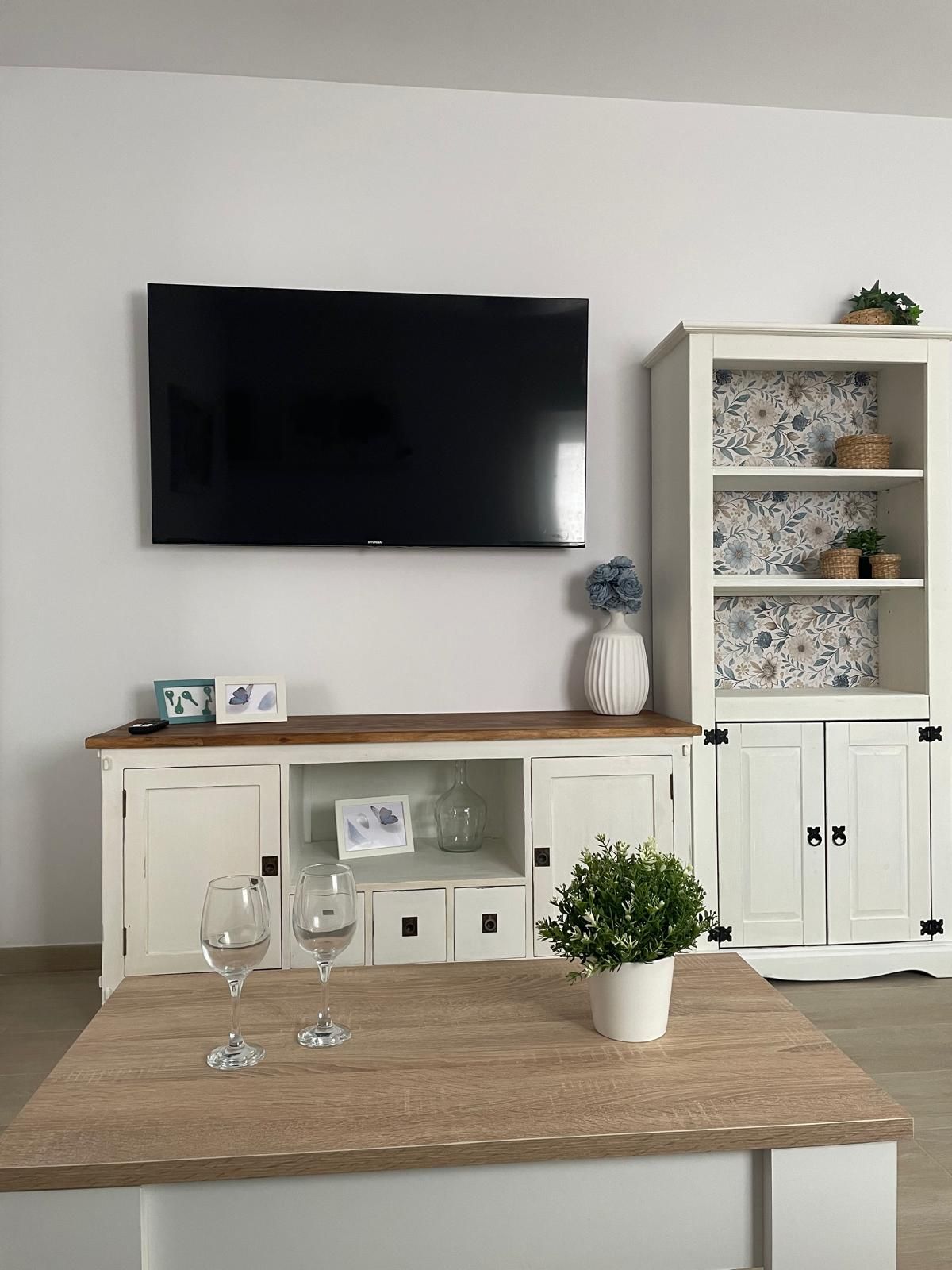 Una sala de estar con un televisor de pantalla plana montado en la pared, una mesa de centro de madera con dos vasos, una planta en maceta y un mueble blanco con papel tapiz floral.