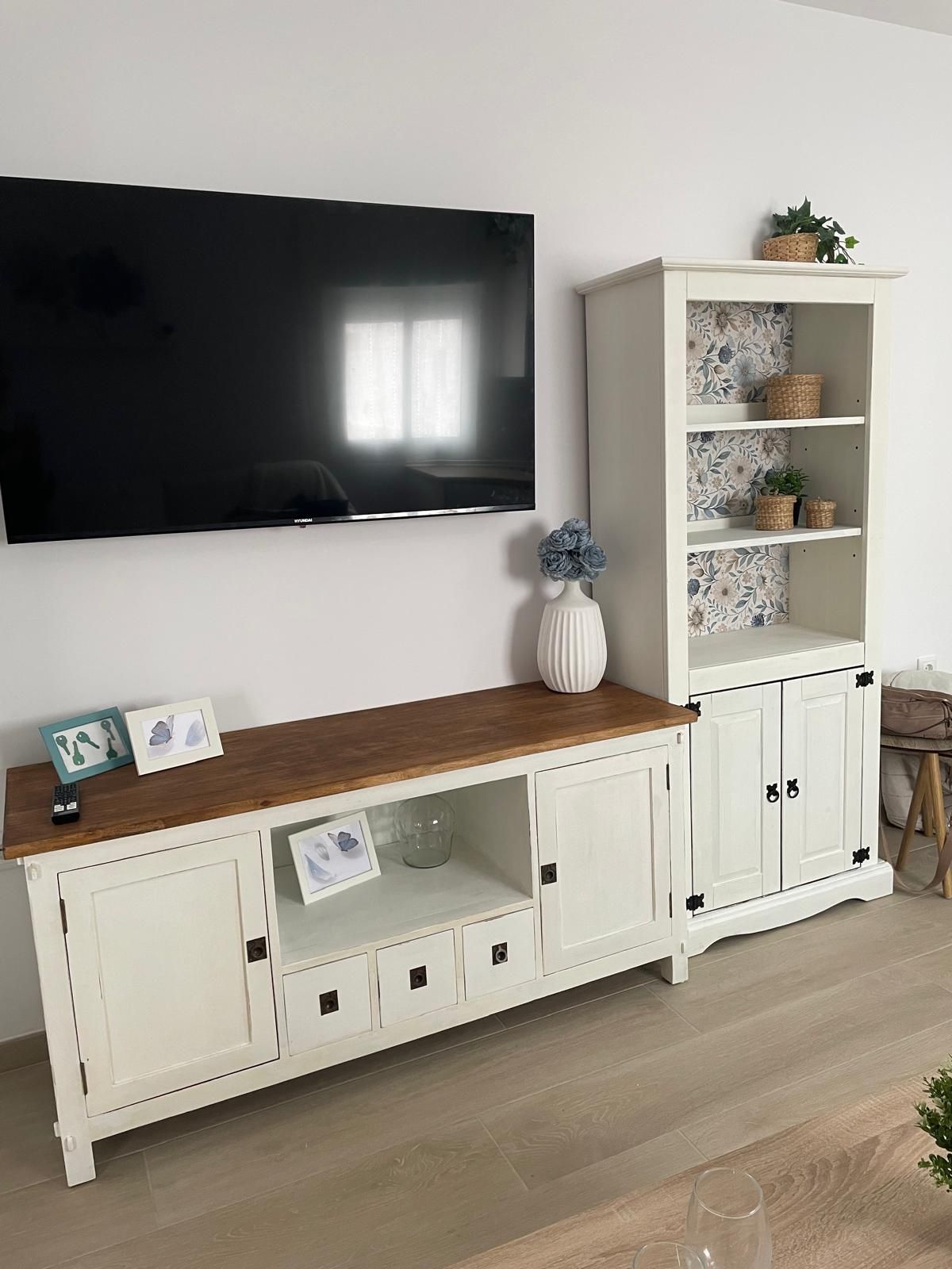 A living room with a flat-screen TV mounted on the wall, a wooden console table with decorative items, and a white cabinet with shelves.