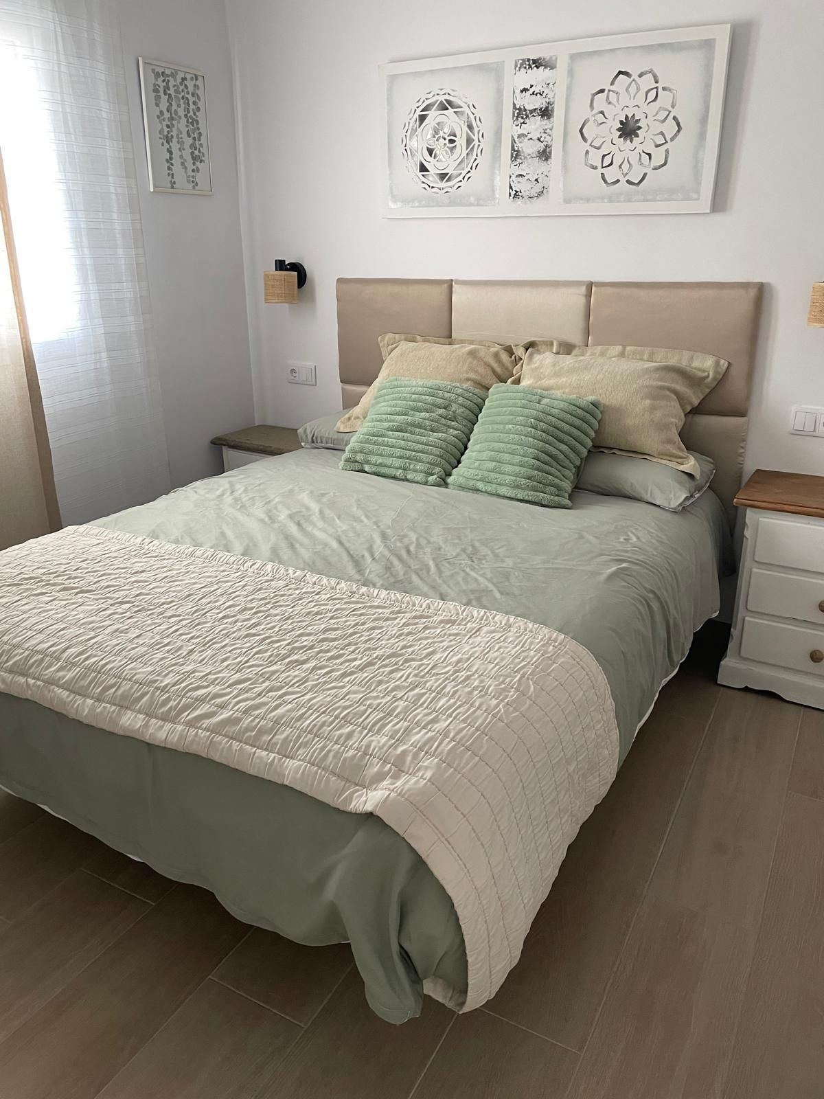 Una cama bien hecha con cubrecolcha acolchado, una mesita de noche con lámpara y cuadros en la pared sobre la cama.