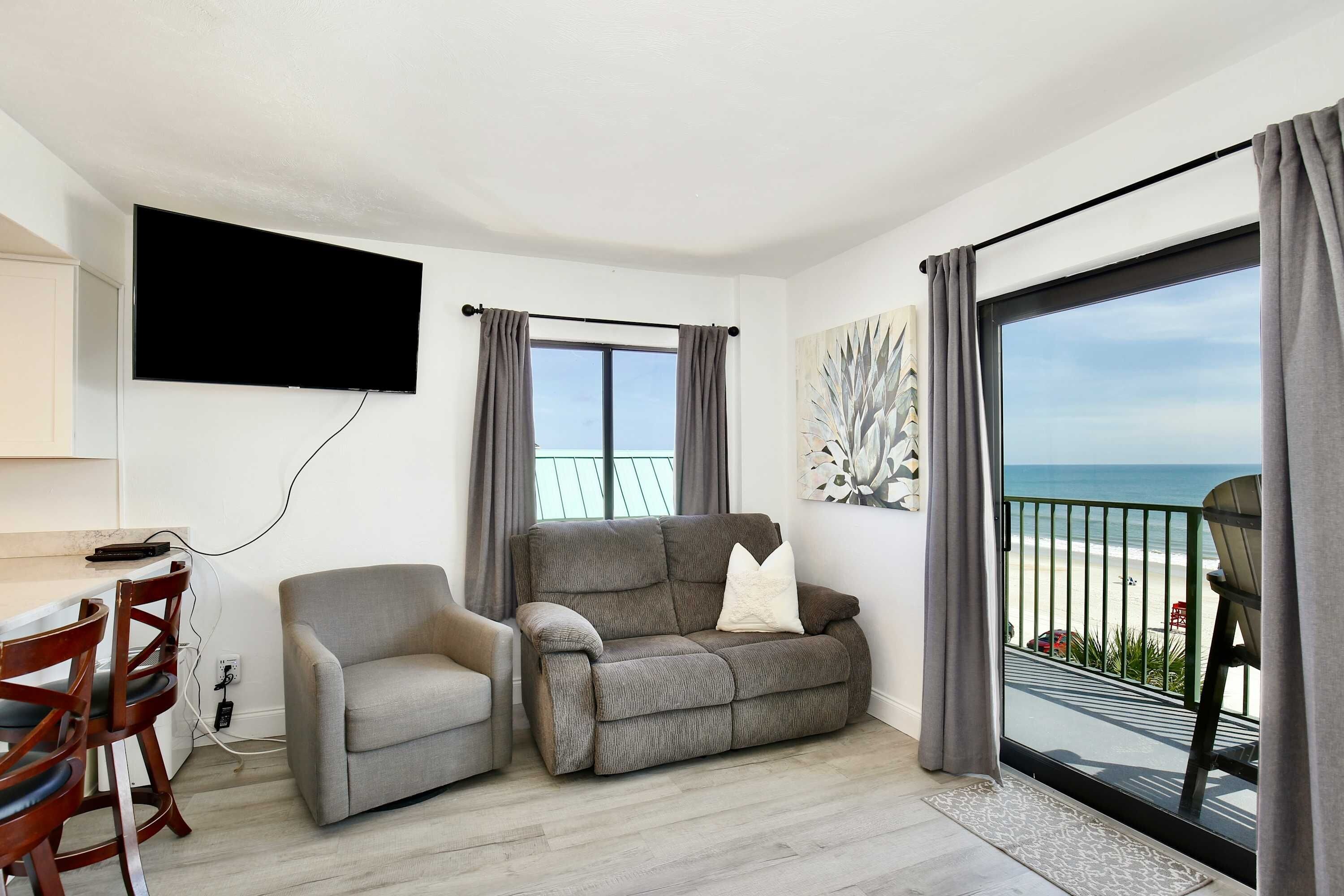 Un salon moderne doté d’un téléviseur à écran plat, d’un canapé gris, d’une chaise en bois et d’un balcon offrant une vue sur la plage.
