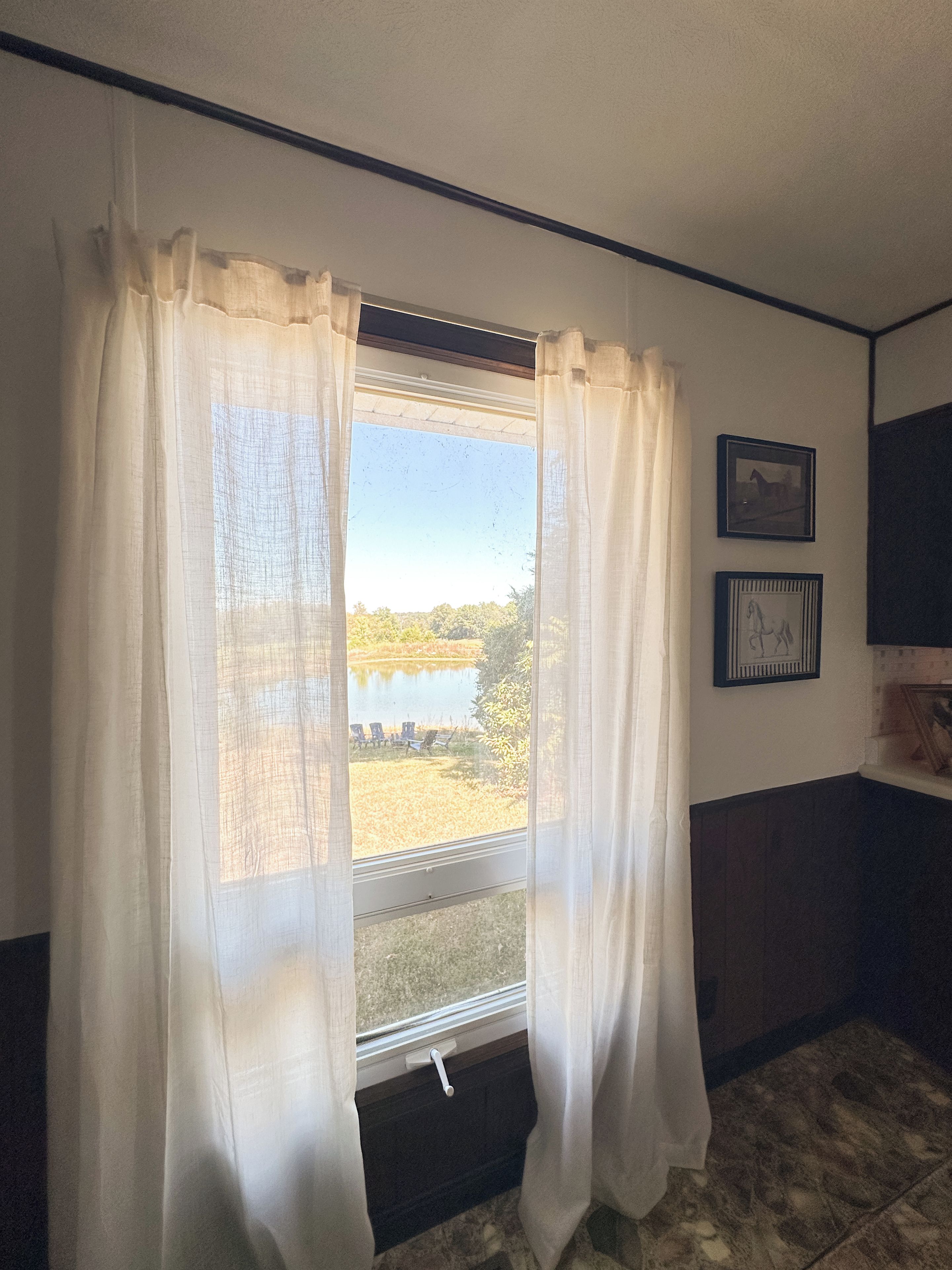 Une pièce avec une fenêtre, des rideaux, un tableau encadré et un sol carrelé.