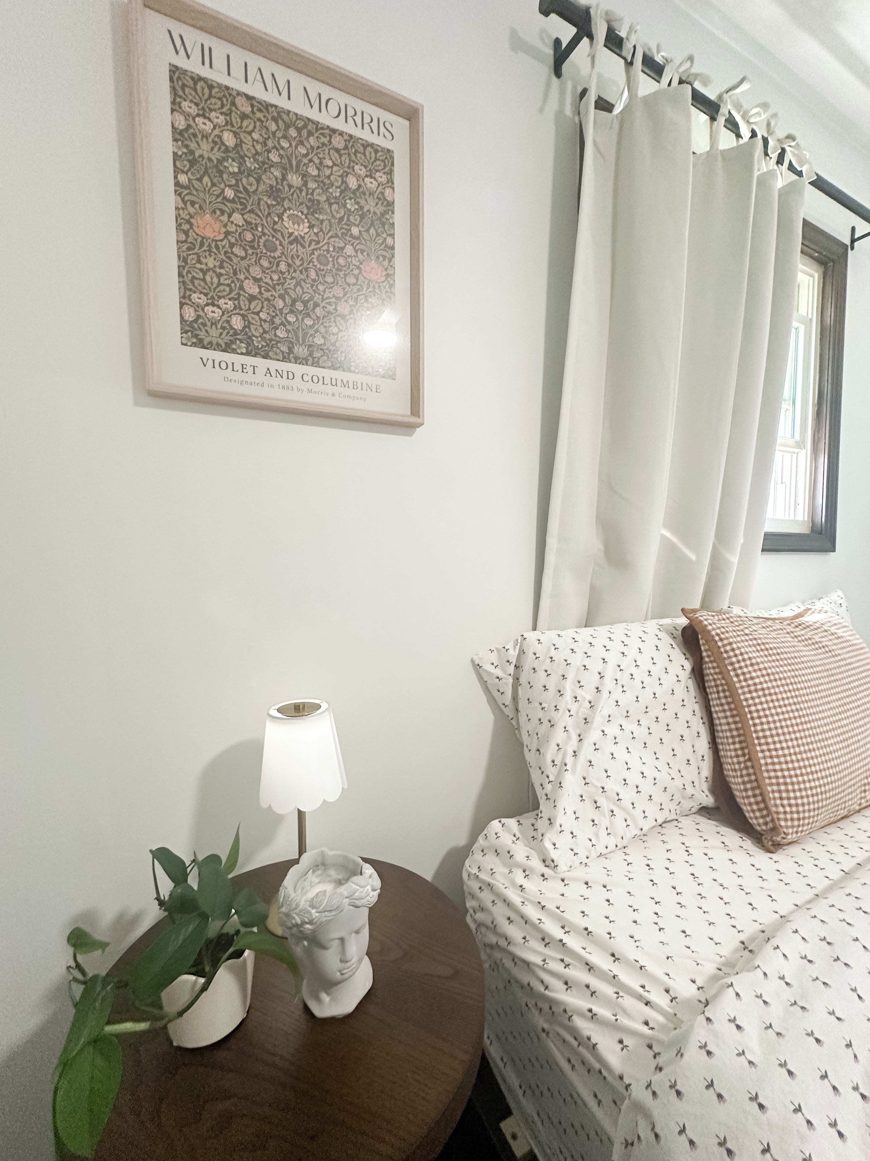 Une pièce comprenant un tableau encadré, une lampe, une plante en pot, une statue, un lit avec une literie à motifs et une fenêtre avec des rideaux.