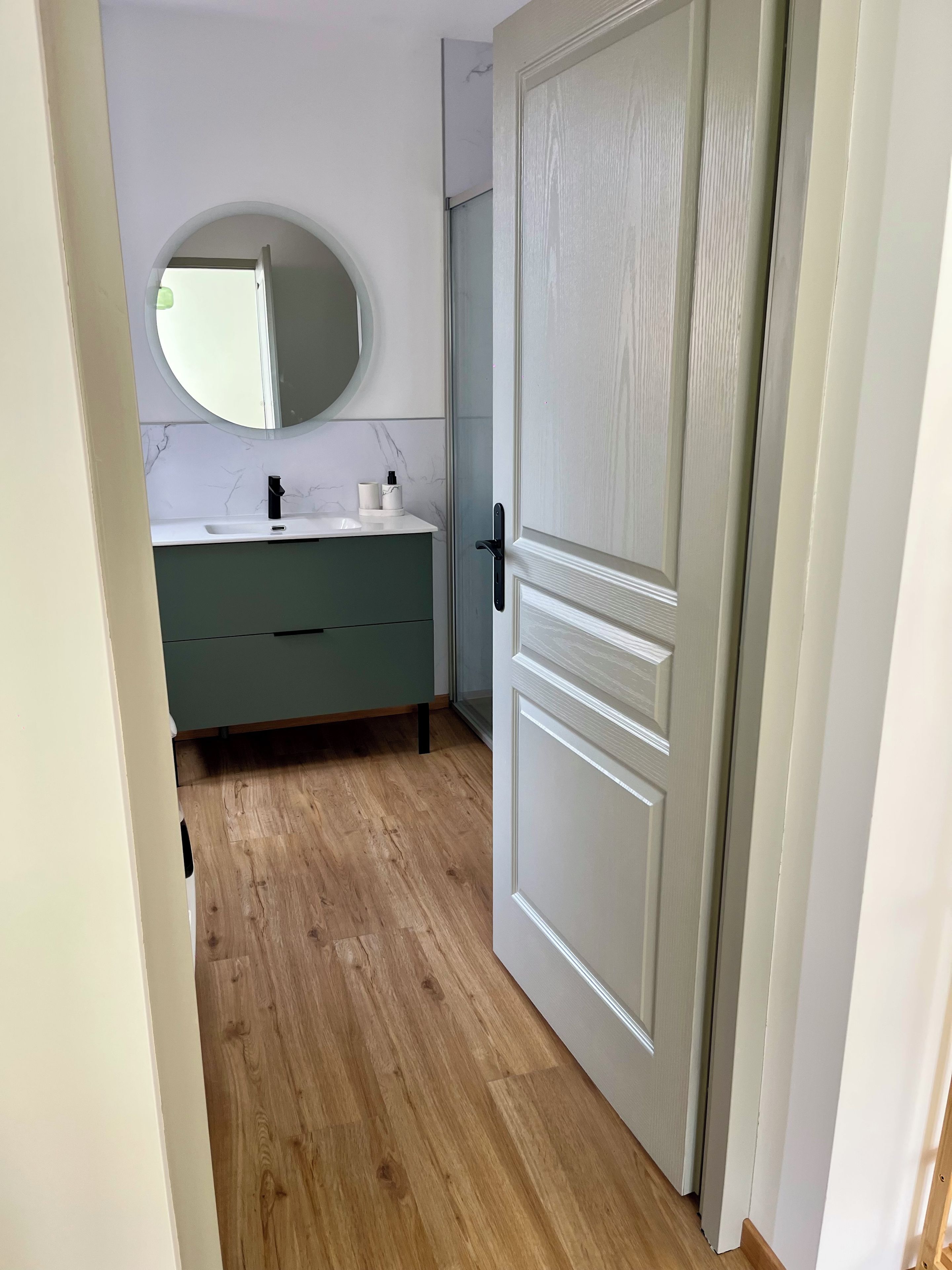 Un pasillo estrecho con piso de madera que lleva a un baño con lavamanos blanco, un mueble verde y un espejo redondo.