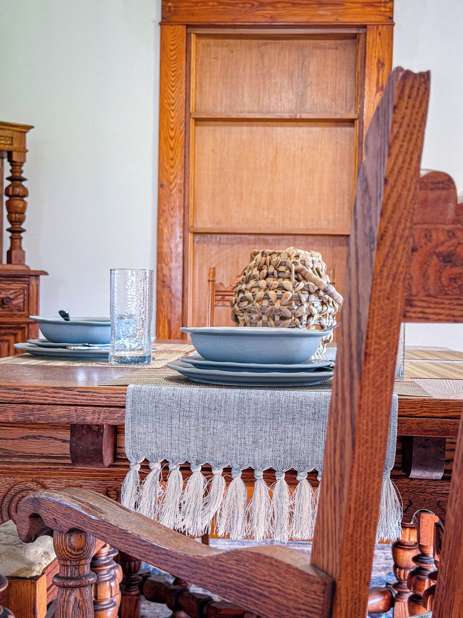 Une table de salle à manger en bois, sur laquelle on a posé un set de table en fibres tressées, un verre et une pile d’assiettes.