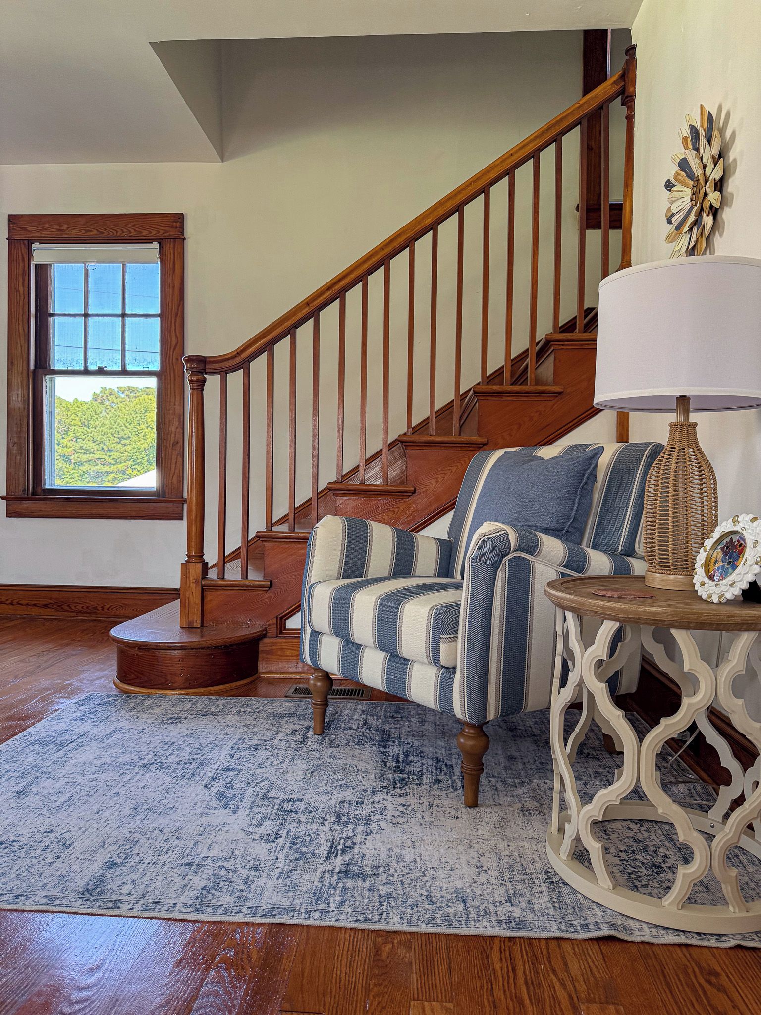 Un salon avec un escalier en bois, un canapé à rayures, un tapis à motifs et une lampe décorative.