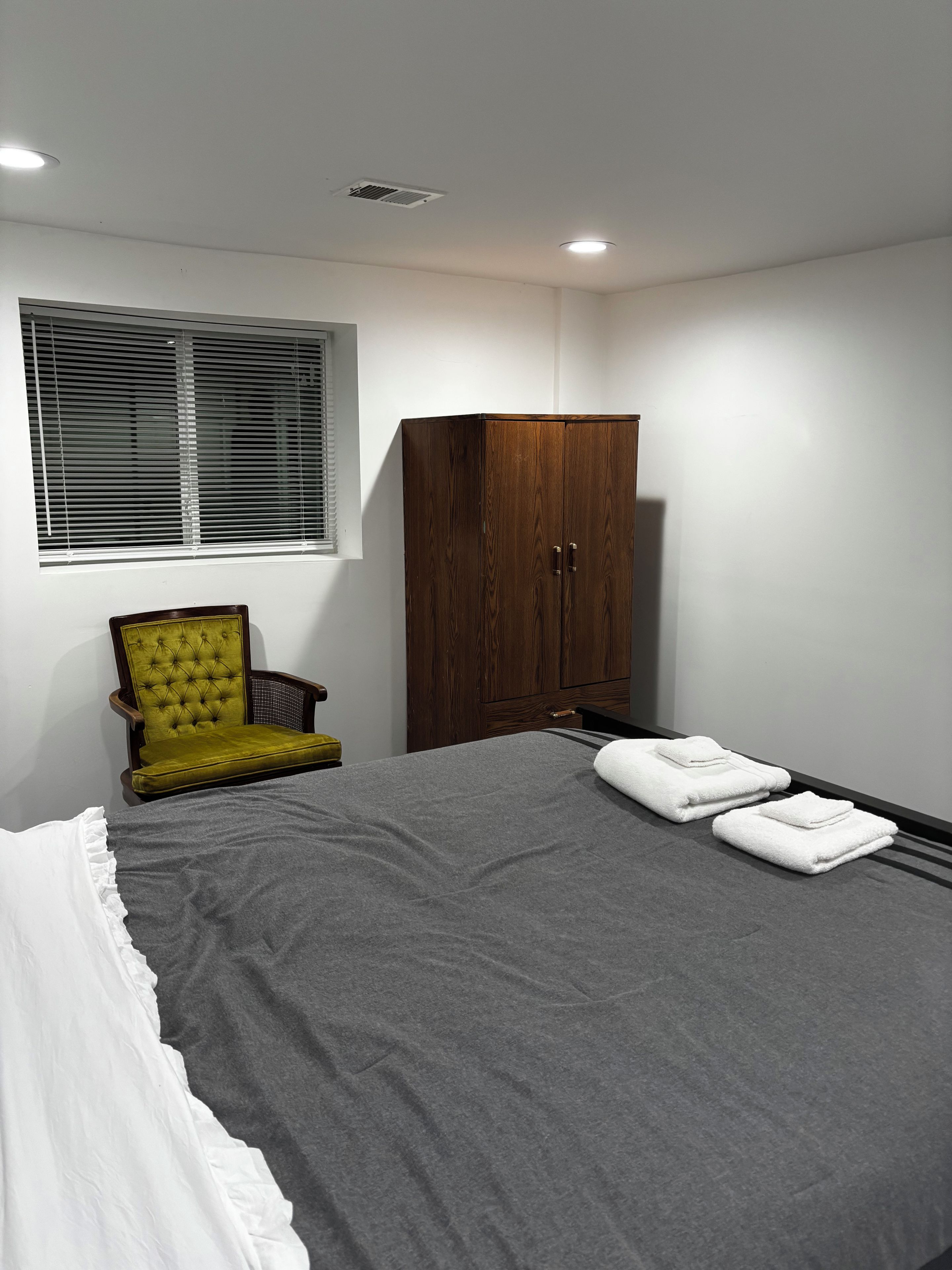 Une chambre à coucher avec un lit, un fauteuil vert, une armoire en bois et une fenêtre avec des stores.