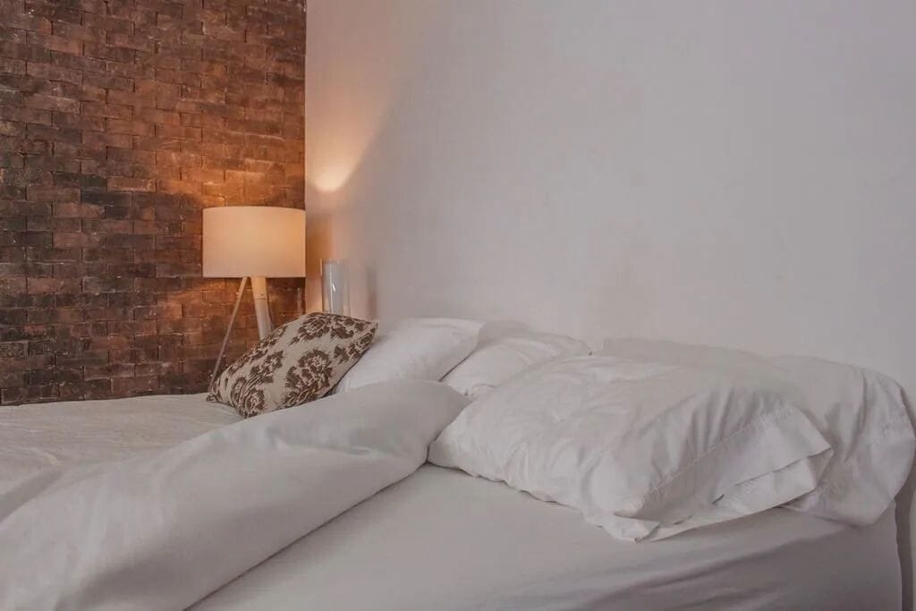 A bed with white bedding, a patterned pillow, and a lamp on a stand.