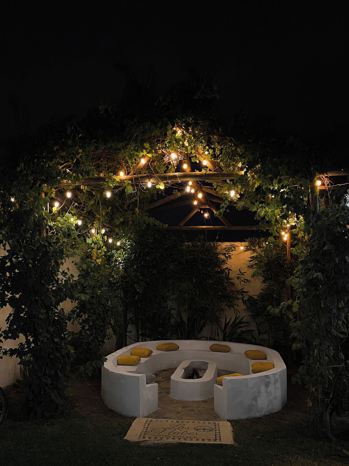 A circular outdoor seating area with a fire pit, surrounded by greenery and string lights.