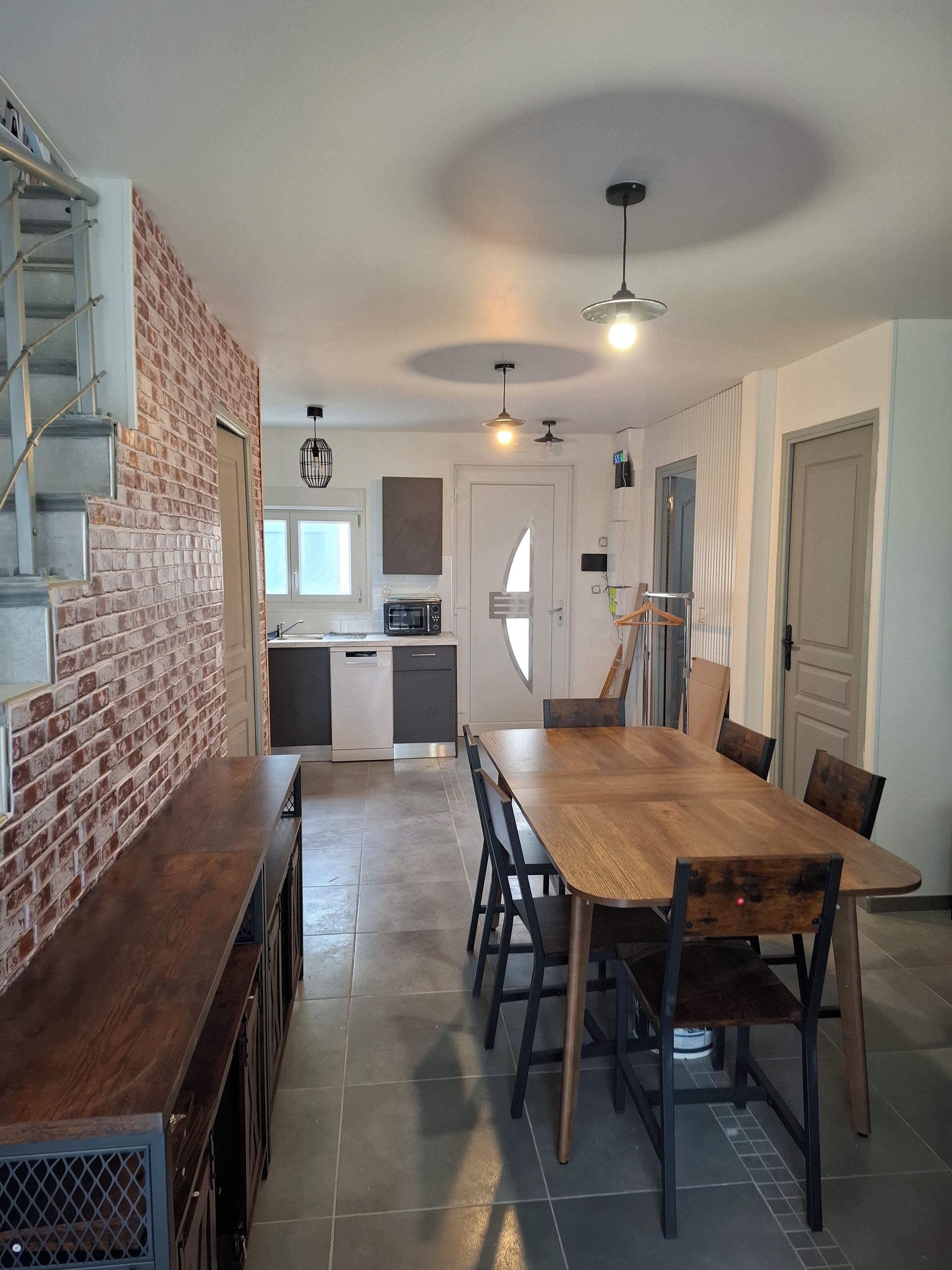 A modern kitchen with a brick accent wall, a dining area with a wooden table and chairs, and a staircase leading to an upper level.