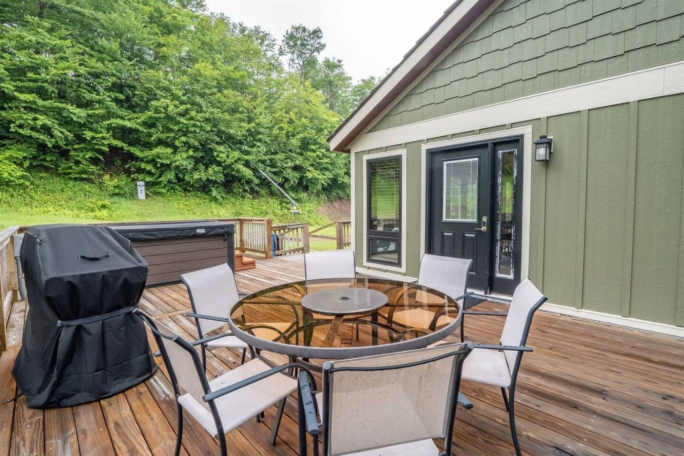 An outdoor deck with a round glass table and chairs, a barbecue grill, and a house with a green exterior.