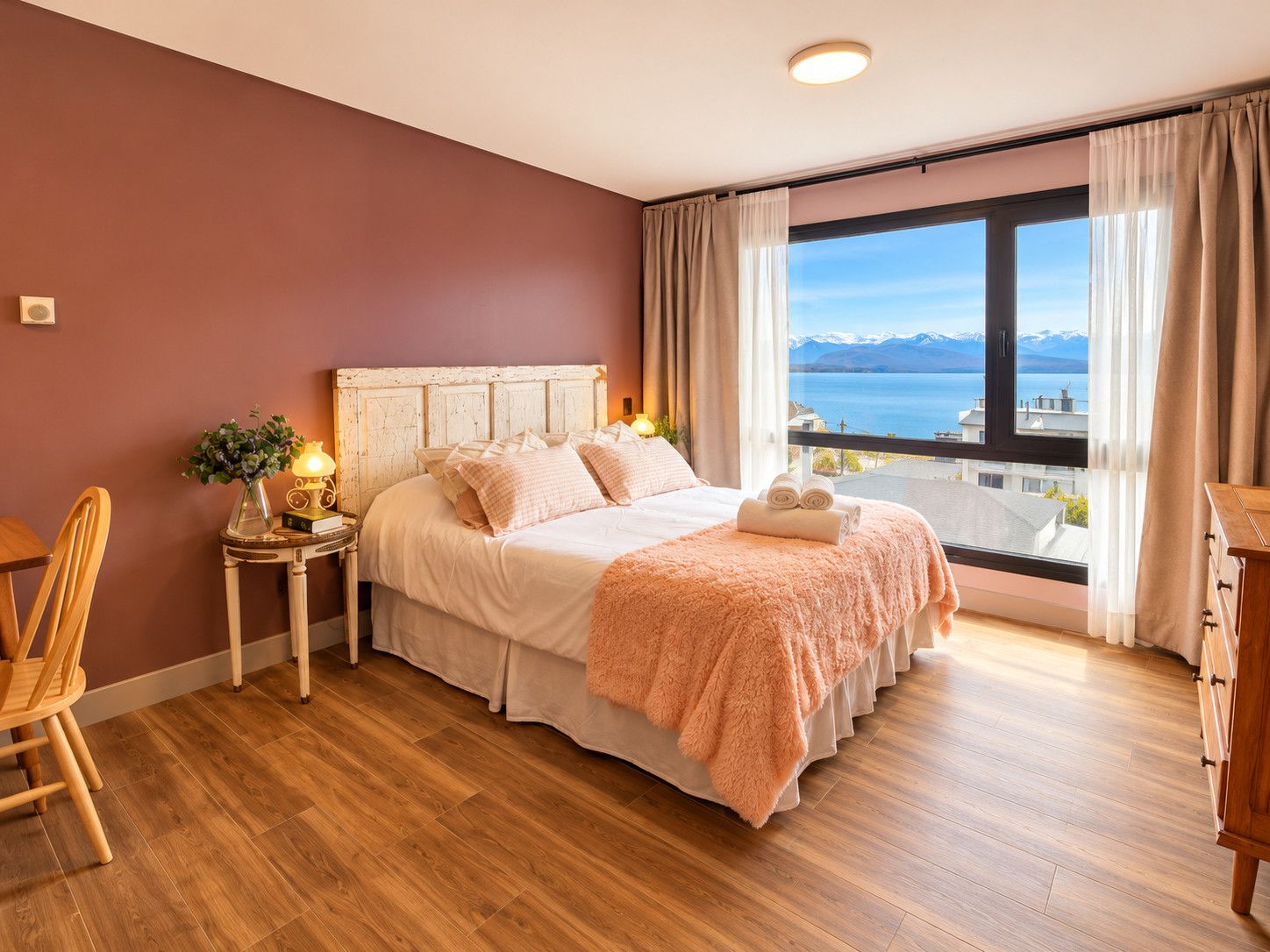 Une chambre avec une grande fenêtre, un lit recouvert d’une couverture rose et moelleuse, une table de chevet avec une lampe, et une vue sur un lac et des montagnes.