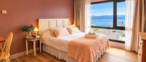 A bedroom with a large window, a bed with a fluffy pink blanket, a bedside table with a lamp, and a view of a lake and mountains.