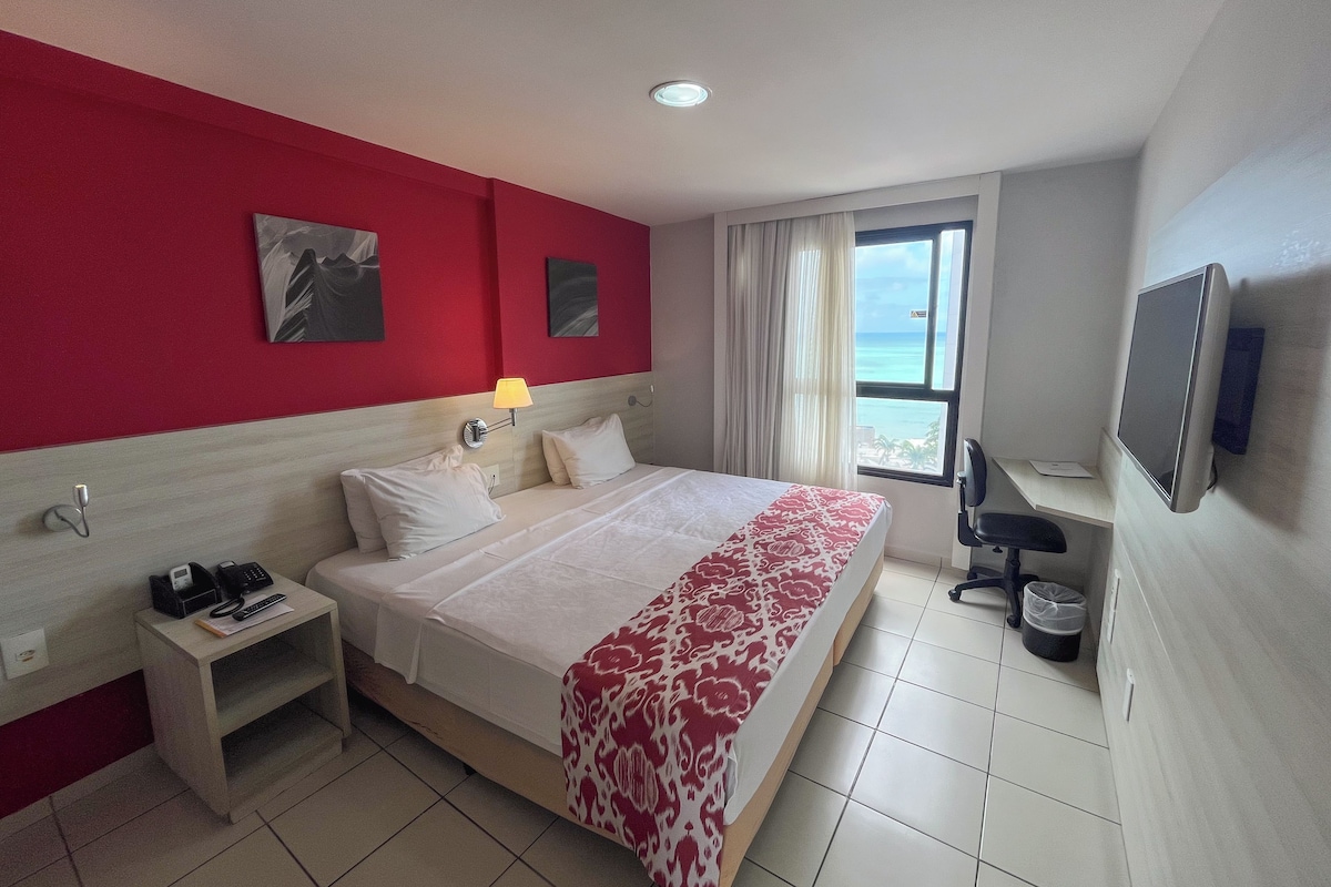 A hotel room with a bed, a desk, a lamp, a TV, and a window with a view of the ocean.