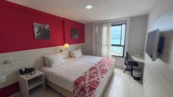 A hotel room with a bed, a desk, a lamp, a TV, and a window with a view of the ocean.
