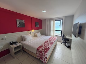 A hotel room with a bed, a desk, a lamp, a TV, and a window with a view of the ocean. - Flats Vista Mar (Natal)
