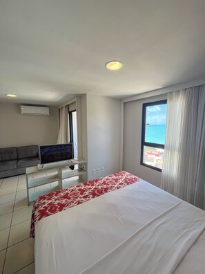 A hotel room with a bed, a sofa, a TV, and a window with a view of the sea. - Flats Vista Mar (Natal)