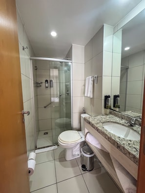 A modern bathroom with a glass-enclosed shower, a toilet, a sink with a granite countertop, and a large mirror. - Flats Vista Mar (Natal)