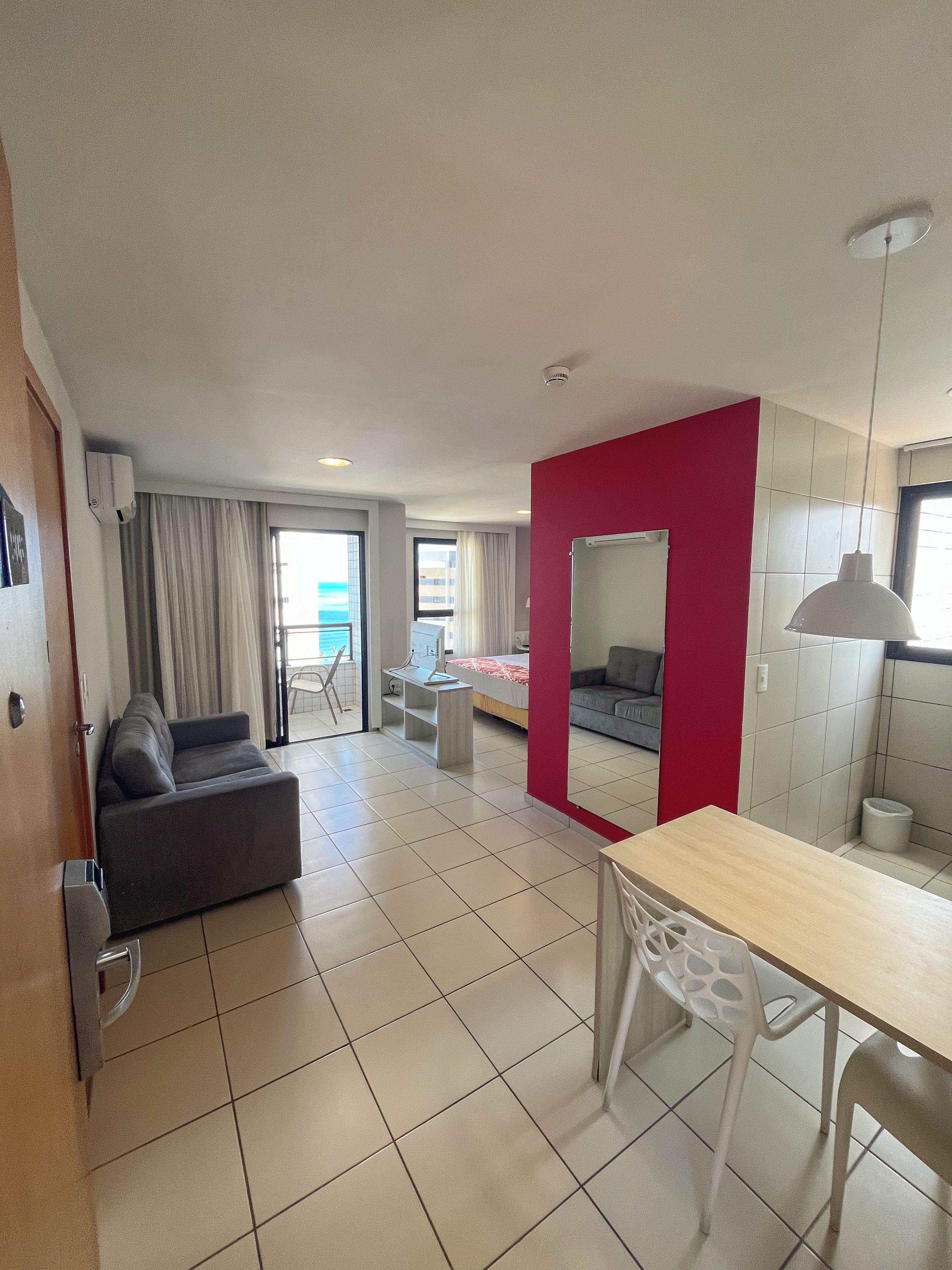 Habitación de hotel moderna con sofá, mesa de comedor con sillas, espejo y vista al mar por la ventana.