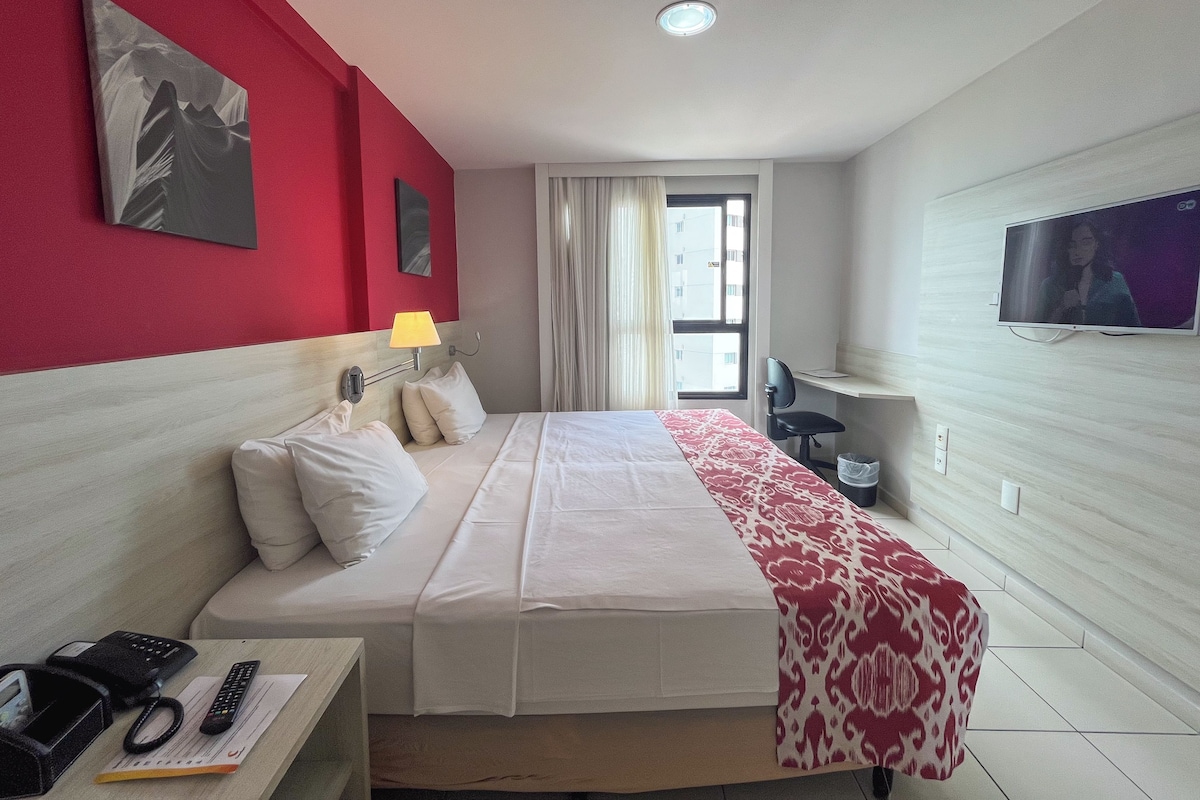 Habitación de hotel con una cama, un escritorio con teléfono y control remoto, un televisor y una ventana con cortinas.